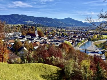 Herbstblick auf Bad Tölz mit Kirchturm, bunten Bäumen, Isar und bayerischen Alpen im Hintergrund unter blauem Himmel.