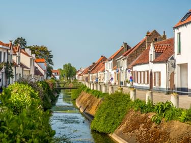 Schmaler Kanal durch Lissewege mit weißen Häusern mit orangeroten Ziegeldächern an beiden Seiten unter klarem blauen Himmel.