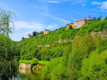 Blick auf die Dornburger Schlösser auf einem grünen Hügel über einem Fluss. Üppige Vegetation bedeckt den Hang, ein kleines Gebäude ist am Wasser sichtbar.