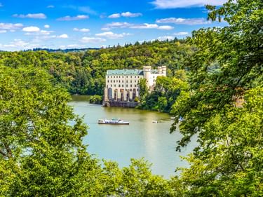 Burg Orlík an der Moldau, umgeben von üppigen grünen Wäldern. Ein Passagierboot fährt auf dem türkisfarbenen Wasser unter blauem Himmel.