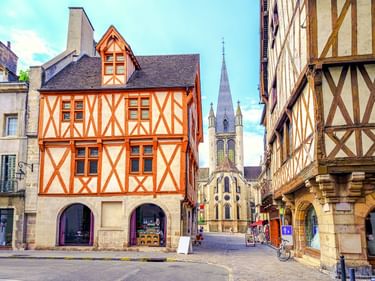 Bunte Fachwerkhäuser mit orangen Balken säumen eine Kopfsteinpflasterstraße in Dijons Altstadt. Ein gotischer Kirchturm ragt im Hintergrund auf.