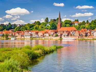 Historische Stadt Lauenburg an der Elbe mit rotgedeckten Häusern und Backsteinkirchturm entlang der Elbe unter blauem Himmel mit weißen Wolken.