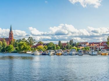 Panoramablick auf Boote am Seehafen mit einem markanten Kirchturm über grünen Bäumen. Blauer Himmel mit weißen Wolken über der Uferpromenade.