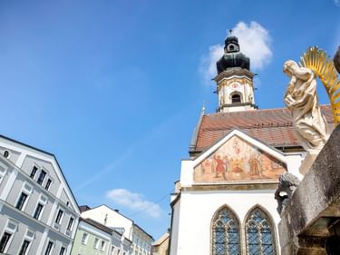 Kirche mit rotem Ziegeldach und Glockenturm in Deggendorfs Altstadt. Eine goldgekrönte Statue steht neben bunten historischen Gebäuden unter blauem Himmel.