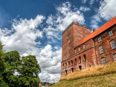 Schloss Koldinghus in Kolding mit roten Backsteinmauern und orangefarbenem Ziegeldach. Dänische Flagge weht vom Turm unter blauem Himmel.