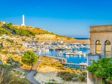 Hafen von Santa Maria di Leuca mit Booten, weißem Leuchtturm auf Hügel, Steingebäude mit Bogenfenstern und Küstenvegetation unter blauem Himmel.
