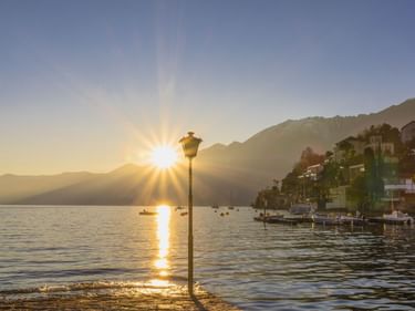 Sonnenuntergang über dem Lago Maggiore mit Bergen, einer Uferstadt, Booten und einer Laterne im Gegenlicht bei Ascona-Locarno.
