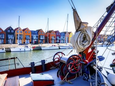 Blick vom Bootsdeck im Hafen Stavoren auf bunte holländische Häuser und vertäute Segelboote. Seile und Segelausrüstung im Vordergrund.