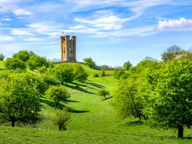 Broadway Tower, ein steinerner Burgturm, steht auf einem grünen Hügel, umgeben von Bäumen unter blauem Himmel mit weißen Wolken.