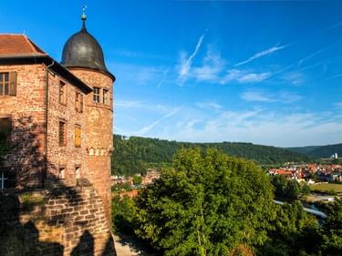 Roter Sandsteinturm der Burg Wertheim mit Kuppeldach mit Blick auf die Stadt Wertheim, umgeben von grünen Wäldern und Hügeln unter blauem Himmel.