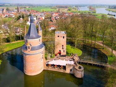Luftaufnahme von Schloss Duurstede mit rundem Turm und Wassergraben, umgeben von Bäumen. Niederländische Stadt mit roten Dächern und Fluss im Hintergrund.