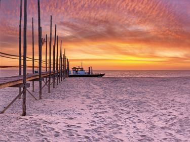 Holzsteg mit Pfählen erstreckt sich ins ruhige Meer bei Sonnenaufgang auf Texel. Orange und rosa Wolken erhellen den Himmel, ein Schiff am Horizont.