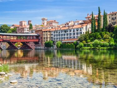 Historische rote überdachte Holzbrücke Ponte Vecchio über einen ruhigen Fluss in Bassano del Grappa mit bunten Gebäuden und Zypressen.