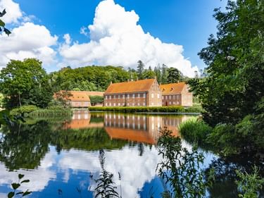 Rotes Backsteinherrenhaus mit orangefarbenem Ziegeldach spiegelt sich in ruhigem See zwischen Vejle und Horsens. Grüne Bäume rahmen die Szene.