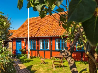 Oranges Fachwerkhaus mit blauen Fenstern und rotem Ziegeldach in Rønne. Garten mit Holzbank und Obstbäumen im Vordergrund unter blauem Himmel.