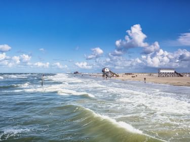 Breiter Sandstrand in St. Peter-Ording mit rollenden Wellen, Pfahlbauten und weißen Wolken über der Nordsee unter blauem Himmel.