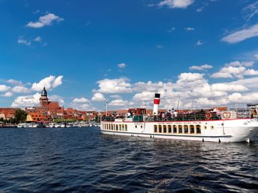 Weißes Flusskreuzfahrtschiff mit rot-schwarzem Schornstein auf blauem Wasser. Historische Stadt mit roten Dächern und Kirchturm unter blauem Himmel.