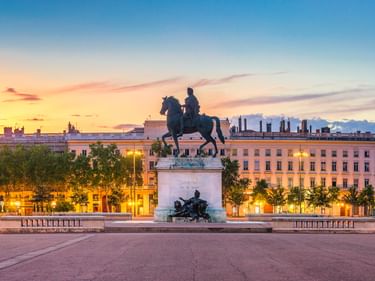 Equestrian statue of Louis XIV on a plaza at sunset, with illuminated classical buildings and colorful sky in the background.