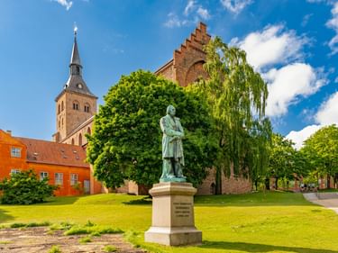 Bronzestatue von H.C. Andersen auf Steinsockel im Park mit rotem Backsteinkirchturm, historischen Gebäuden und grünem Rasen in Odense.