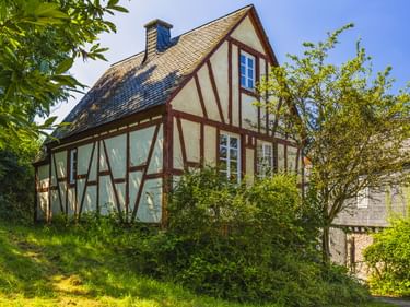 Traditionelles Fachwerkhaus mit braunen Holzbalken und cremefarbenen Wänden in Konz, umgeben von grünen Bäumen und einem Hanggarten unter blauem Himmel.