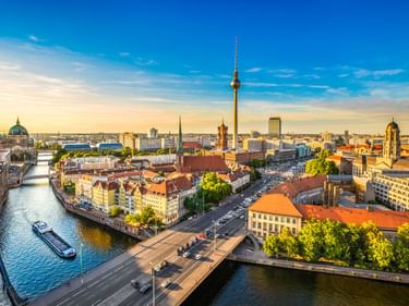 Luftaufnahme von Berlin bei Sonnenuntergang mit Fernsehturm, Spree mit Boot, historischen Gebäuden und Brücken unter blau-goldenem Himmel.