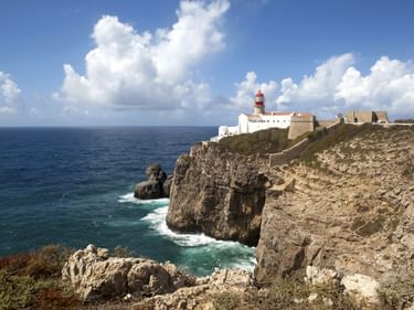 Rot-weißer Leuchtturm am Cabo de São Vicente auf dramatischen Klippen mit Blick auf den Atlantik und weißen Wolken am blauen Himmel.