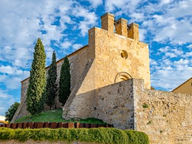 Pfarrkirche Sant Vicenç in Vilamalla mit Steinfassade, drei Turmzinnen und Rundfenster. Zypressen und Hecken rahmen das Gebäude ein.
