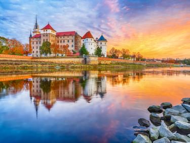 Schloss Hartenfels mit bunten Türmen spiegelt sich bei Sonnenuntergang in der Elbe. Herbstbäume säumen das Flussufer unter dramatischem Himmel.