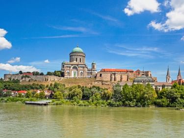 Basilika Esztergom mit grüner Kuppel auf einem Hügel mit Blick auf die Donau, umgeben von historischen Gebäuden und üppigem Grün unter blauem Himmel.