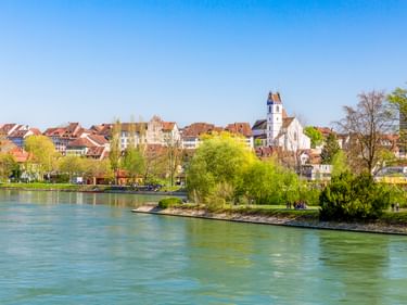Blick auf Aarauer Altstadt mit Kirchturm über die Aare. Grüne Bäume säumen das Flussufer unter klarem blauen Himmel in der Schweiz.