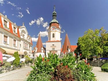 Historic Steiner Tor gate with white tower and green dome in Krems. Flower beds and trees in foreground, red-roofed buildings on left, blue sky.
