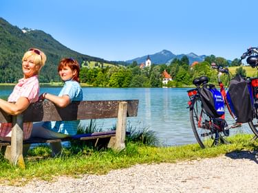 Zwei Frauen sitzen auf Holzbank am türkisfarbenen See mit Fahrrädern in der Nähe. Berglandschaft und Kirche im Hintergrund bei Füssen.