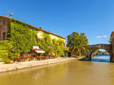 Canal du Midi mit efeubedeckten Steingebäuden entlang des Wasserwegs. Ein Steinbrückenbogen rahmt die Ansicht, Palmen und blauer Himmel sind sichtbar.