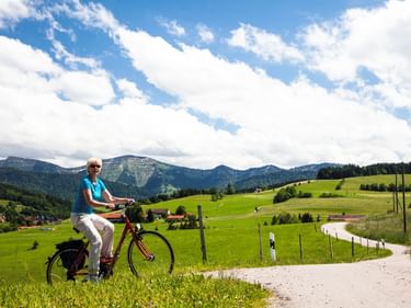 Frau in blauem Shirt radelt auf Schotterweg durch grüne Wiesen im Allgäu, Bayern. Berge und Dorf im Hintergrund unter bewölktem Himmel.