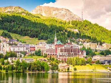Blick auf St. Moritz mit bunten Gebäuden am See, bewaldeten Hängen und Bergen mit Sendeturm unter teilweise bewölktem Himmel.