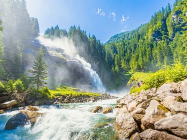 Krimmler Wasserfälle stürzen bewaldete Berghänge hinab, Nebel steigt über dem rauschenden Wasser auf, das über felsiges Gelände fließt.