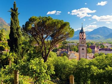 Blick auf Meran mit einem markanten Kirchturm inmitten üppiger Vegetation. Berge und blauer Himmel mit Wolken rahmen die italienische Stadt.