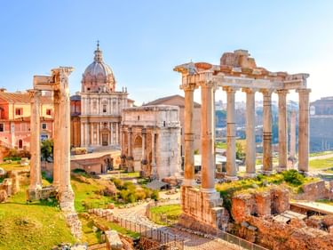 Antike römische Säulen und Ruinen am Forum Romanum mit barocker Kirchenkuppel und historischen Gebäuden im Hintergrund unter blauem Himmel.