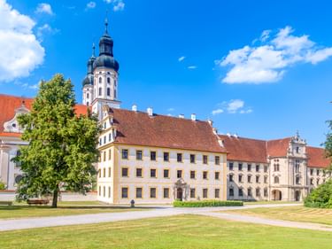 Obermarchtal Monastery complex with white baroque church towers, red-tiled roofs, and yellow facade buildings surrounded by green lawns and trees.