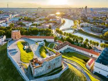 Aerial view of Vilnius showing a historic fortress with round tower, river Neris winding through the city, and modern skyline at sunset.