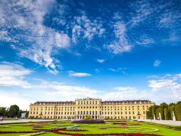Yellow Baroque palace with ornamental gardens featuring geometric flower beds in red, white, and green under a blue sky with white clouds.
