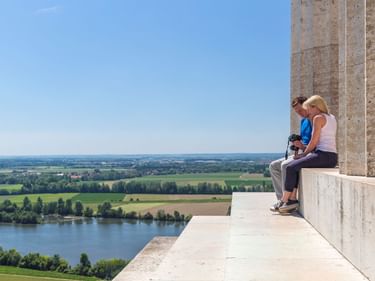 Zwei Personen sitzen auf der Steinbrüstung der Walhalla und blicken auf die Donau und die grüne Tallandschaft unter blauem Himmel.