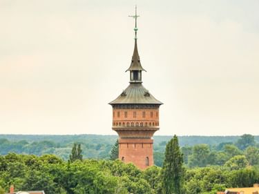 Red brick water tower in Forst with green copper roof and spire, rising above surrounding trees against a pale sky.