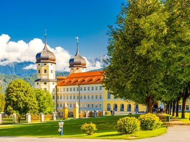 Stift Stams Kloster mit gelber Fassade, orangefarbenem Dach und zwei Zwiebeltürmen. Grüne Rasenfläche und von Bäumen gesäumter Weg im Vordergrund, Berge dahinter.