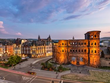 Das antike römische Stadttor Porta Nigra bei Abenddämmerung beleuchtet in Trier, mit historischen Gebäuden und farbigem Himmel.