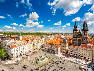 Luftaufnahme des Altstädter Rings in Prag mit den gotischen Türmen der Teynkirche, bunten historischen Gebäuden und Besuchern unter blauem Himmel.