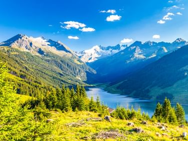 Panoramablick vom Gerlospass auf einen blauen Bergsee, umgeben von bewaldeten Hängen und schneebedeckten Alpengipfeln unter hellem Himmel.