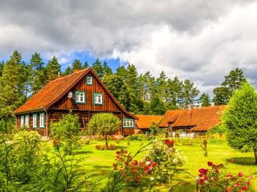 Holzbauernhaus mit orangefarbenem Ziegeldach in Krutyn, umgeben von grünem Rasen, blühenden Rosen und Wald unter dramatischem Wolkenhimmel.