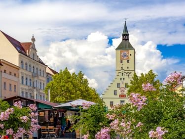 Historic clock tower in Deggendorf old town with colorful buildings, pink flowering bushes, and outdoor café under blue sky with clouds.