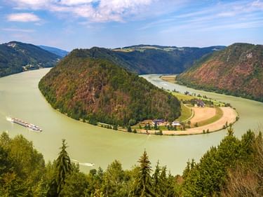 Luftaufnahme der Donauschlinge bei Schlögen, die eine Hufeisenkurve um eine bewaldete Halbinsel mit Feldern und Häusern bildet, Frachtschiff auf dem Wasser.